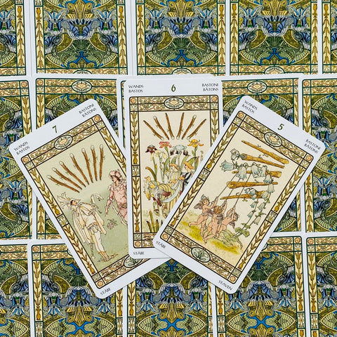 Tarot kortos su baltu rėmeliu, fanuotos trys Lazdų kortos (5–7) centre, padėtos ant mėlyno‑žalsvo ornamentuoto nugarėlių fono