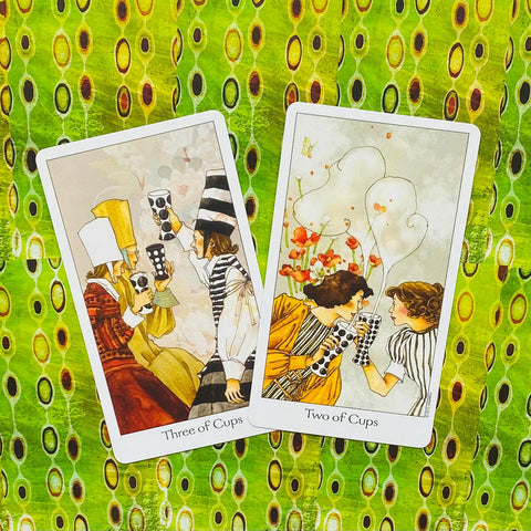 tarot kortos, atverstos dvi kortos (Two of Cups ir Three of Cups) ant žalsvo raštuoto fono, demonstruoja kortų iliustracijų variantą
