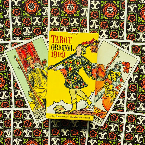 Tarot kortos Original 1909 geltona dėžutė su klajūno iliustracija, padėtos ant išskleistų kortų ir ornamentuoto nugarėlių rašto