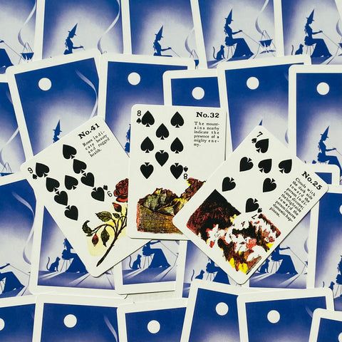 Lenormand kortos su mėlynais nugarėliais, trys atverstos figūrinės kortos (rožė, kalnai, ugnis) parodo konkretaus varianto detalę