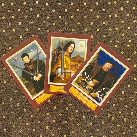 Tarot kortos su rudais rėmeliais, trys atverstos kortos centre vaizduoja kardus ir taurę ant tamsaus raštuoto audinio