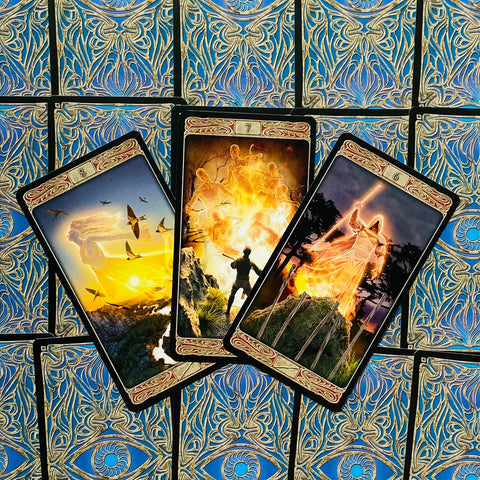 tarot kortos stalo dizaine: trys atvertos kortos su ugnies motyvais ir mėlynomis auksuotomis nugarėlėmis, persidengiančios kampu