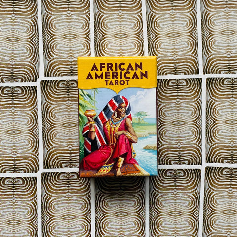Tarot kortos African American Tarot: kompaktiška geltono viršelio dėžutė su spalvota figūrine iliustracija, centre ant tekstūruoto nugarėlių fono
