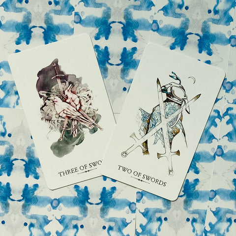 Tarot kortos su Two of Swords ir Three of Swords akvareliniais piešiniais ant mėlyno raštuoto fono