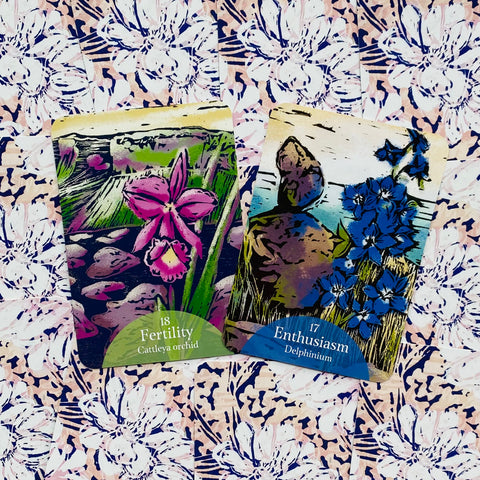 Orakulų kortos su ryškiomis gėlių iliustracijomis — rožinė Cattleya (Fertility) ir mėlyna Delphinium (Enthusiasm) ant pastelinio raštuoto fono