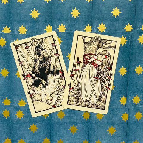tarot kortos ant mėlyno fono su auksinėmis žvaigždėmis, dvi kreminės kortos su raudonais kardų akcentais ir skirtingomis iliustracijomis