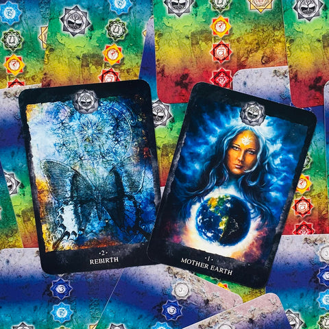 Orakulų kortos su dviem priekiniais paveikslais – "Mother Earth" ir "Rebirth" mėlynai‑spalvotame čakrų fone, plokščiai išdėstytos kampu