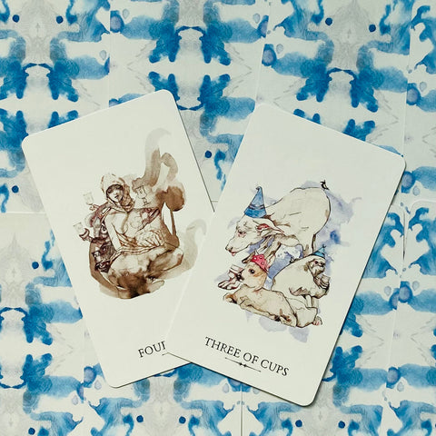 Tarot kortos su akvarelinėmis iliustracijomis, dvi išklotos kortos (Three of Cups ir Four), pakreiptos ant mėlyno raštuoto nugarėlių fono