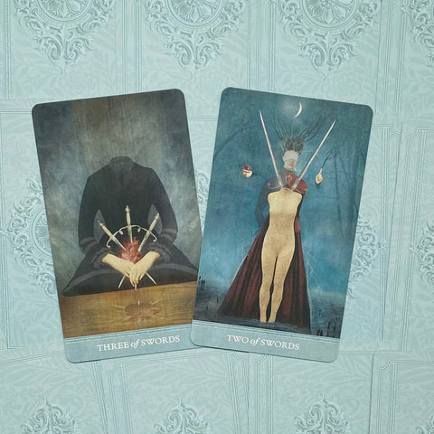 Tarot kortos komplektas: dvi kortos (Three of Swords, Two of Swords) ant turkio fono, kampu nusuktos, akcentuojančios kardų motyvus ir skirtingus paveikslų dizainus