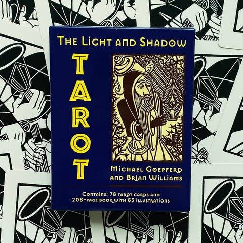 The Light and Shadow Tarot mėlynas dėklas su geltonu užrašu ir linorėžinio stiliaus iliustracija priekyje, fotografuota iš priekio ant juodai baltų kortų fono