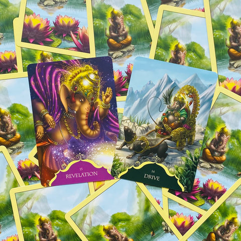 Orakulų kortos su Ganesha iliustracijomis — atviros Revelation (purpurinė) ir Drive (mėlyna) kortos ant geltonų nugarėlių
