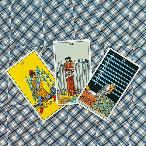 tarot kortos Rider‑Waite: trys Kalavijų kortos išdėstytos fanuote ant mėlyno raštuoto fono, matomas kortų priekis ir iliustracijos