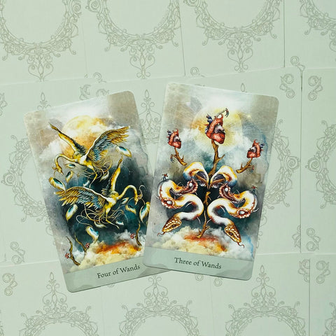 Tarot kortos, dvi atviros kortos (Four of Wands ir Three of Wands) su auksinėmis ir mėlynomis iliustracijomis, parodytos iš viršaus ir fone ornamentinis raštas