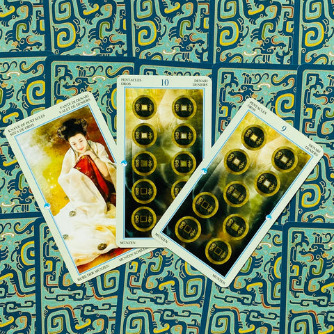 Tarot kortos trijų kortų priekinis išdėstymas (Pentaklių rinkinys: 9, 10 ir rūmų korta) ant mėlyno ornamentuoto fono, artimas planas