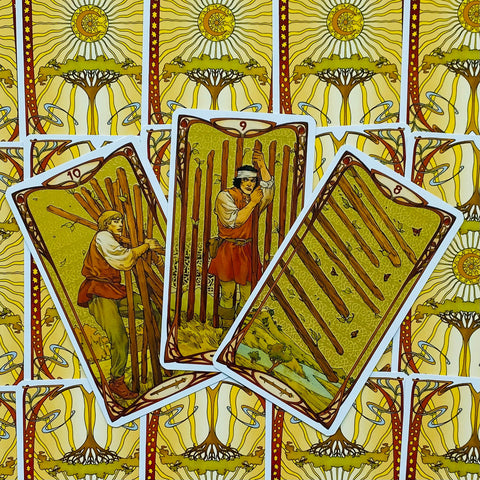 Tarot kortos su aukso geltonu nugarėlių raštu, trys atverstos kortos fanuotos, išryškinančios lazdų simboliką ir detalų dizainą