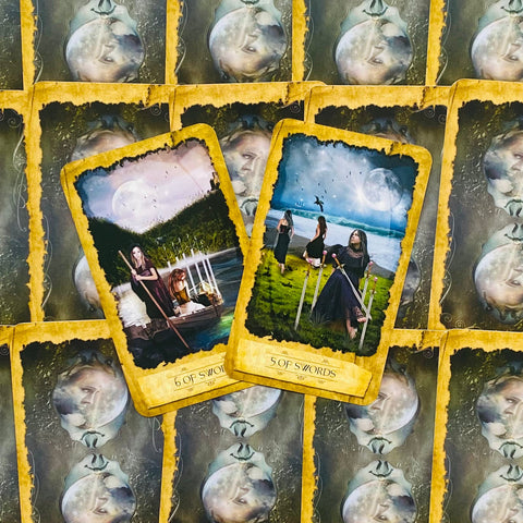 tarot kortos su geltonais kraštais: matomos 5 ir 6 kalavijų kortos prie mėlyno mėnulio, kortų nugarėlės fone