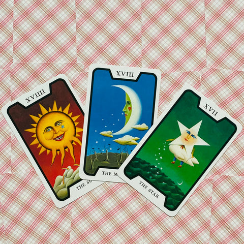 Tarot kortos trijų arkana: Saulė (geltona/raudona), Mėnulis (mėlyna) ir Žvaigždė (žalia) gulsčiai išdėstytos ant rožinio languoto fono.