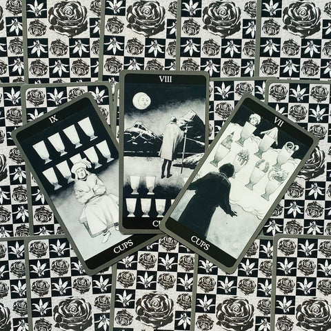 Tarot kortos juodai baltos — trijų Taurių kortų išdėstymas iš viršaus ant rožių ir lapų raštuoto fono, pabrėžia simboliką