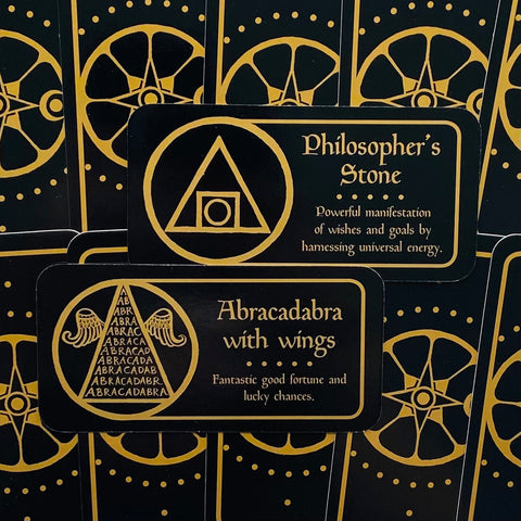 Orakulų kortos juodos su auksinėmis detalėmis, artimas priekinis kadras, rodantis Philosopher's Stone ir Abracadabra dizainus