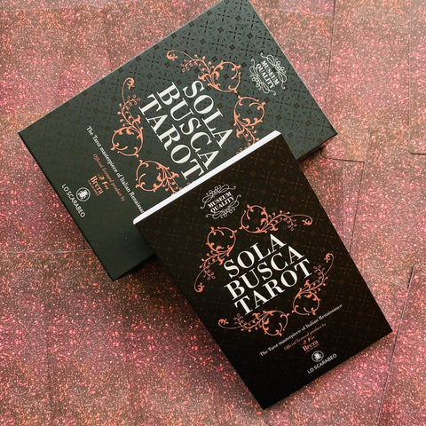 Sola Busca tarot dėžutės (juodos su rožinio aukso ornamentais), dvi skersai išdėstytos ant rausvo blizgaus, blizgančio fono