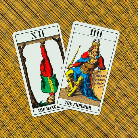 Tarot kortos Rider–Waite stiliaus: dvi kortos — XII Pakabintasis ir IV Imperatorius ant geltono raštuoto fono, kampinis artimas kadras