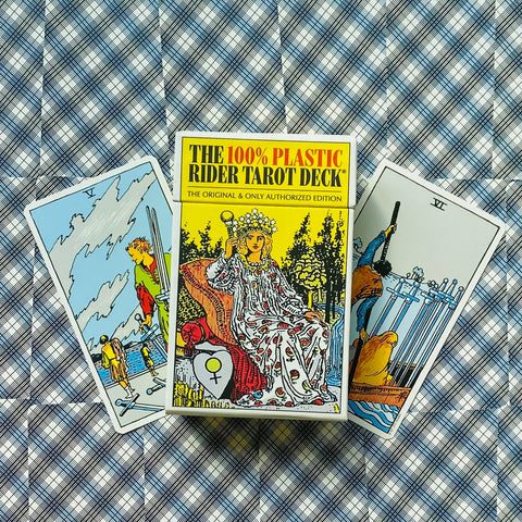 Rider Tarot 100% Plastic geltona pakuotė centre su dviem išskleistomis kortomis šone, viršutinis kampuotas planas