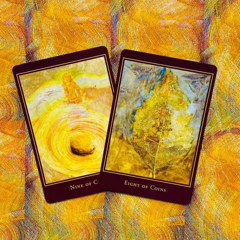 Tarot kortos atvertos – auksinės geltonos devyneto ir aštuoneto monetų kortos ant raštuoto geltono fono, pabrėžiančios kaladės meninį stilių