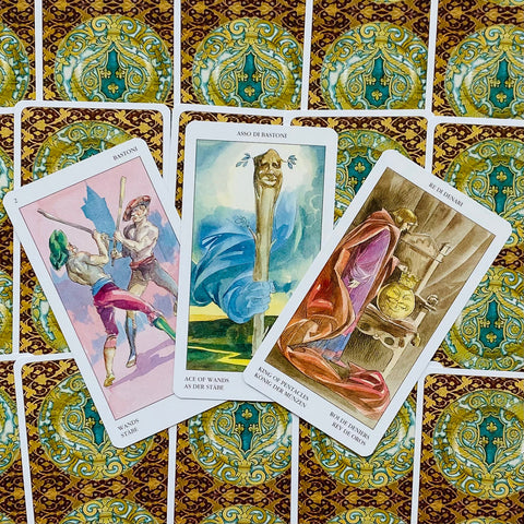 Tarot kortos su auksiniu–žaliu nugarėlių raštu, trys atverstos kortos (Asas lazdučių, dvejetukas lazdomis, Pentaklių karalius)