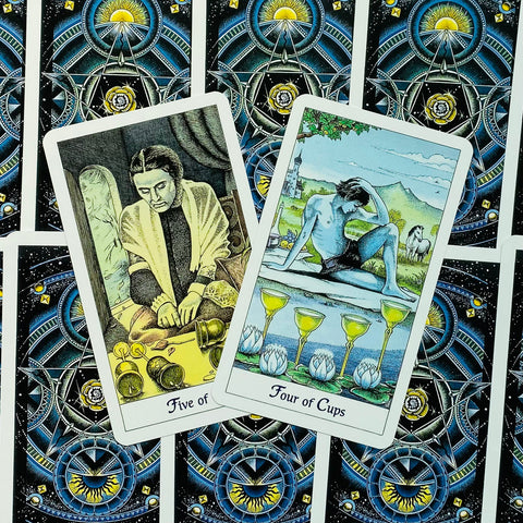 Tarot kortos Rider–Waite stiliaus, dvi atverstos kortos – Keturių ir Penkių taurų motyvai ant mėlynos kosminės nugarėlės, skirtingas kampas