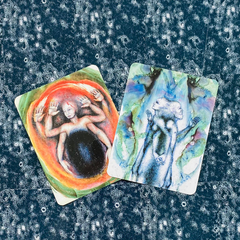 tarot kortos komplektas: dvi iliustruotos kortos, viena oranžinė su juodu centru, kita mėlyna su veidų duetais, persidengiančios ant raštuoto fono