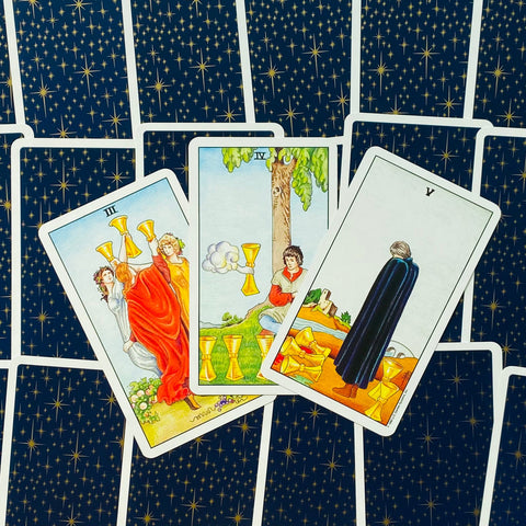 Tarot kortos su mėlynomis žvaigždėtomis nugarėlėmis, trys Taurių rūšies kortos atverstos ir išdėstytos kampu, stalinis stiliaus renginys