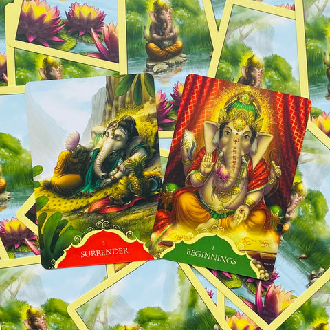 Orakulės kortos su spalvingais Ganesha iliustracijomis, dvi atverstos kortos „Surrender“ ir „Beginnings“, artimas kampas, blizgus laminuotas paviršius
