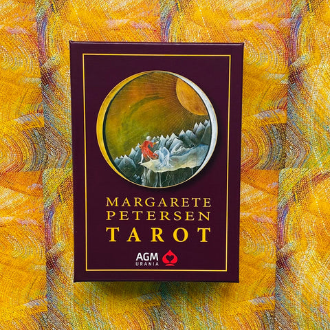 Tarot kortos Margarete Petersen — bordo dėžutė su aukso užrašu ir apskritu iliustracijos langu ant geltono raštuoto fono