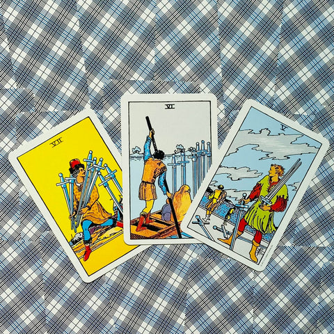 tarot kortos Rider‑Waite stiliaus, trys kardų kortos (VII, VI, V) ant mėlyno‑baltos languotos staltiesės, artimas plokščias išdėstymas