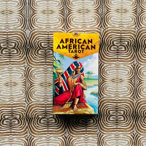 tarot kortos African American Tarot dėžutė su geltonu viršeliu ir spalvota moters iliustracija, fotografuota iš viršaus ant raštuoto rudo fono