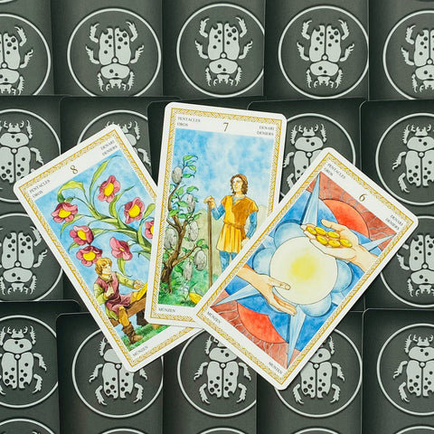 Tarot kortos trys atverstos Monetų rūšies kortos su spalvingomis akvarelinėmis iliustracijomis ant juodų vabalų nugarėlių, skirtos būrimui