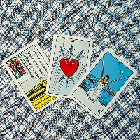 Tarot kortos Rider–Waite stiliaus, trijų kortų išklotinė su Trejais kardais centre, ryškūs raudoni ir mėlyni tonai ant languoto fono