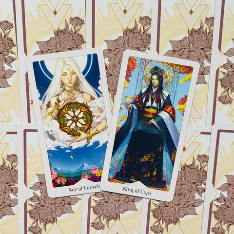 Tarot kortos su iliustracijomis — dvi atverstos kortos (Ace of Laurels ir King of Cups) ant dekoratyvaus galinio rašto, skirtos produkto meno rodymui