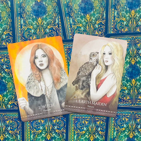 Orakulo kortos su atverstu priekiniu dizainu: oranžinė Witch (Aistra) ir smėlio Earth Maiden (Gamta) ant mėlyno ornamentinio fono