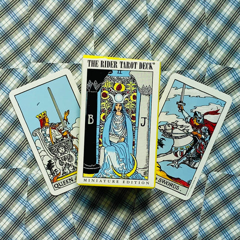 Rider tarot kortos mini dėžutė su geltonu kraštu, padėta tarp dviejų išskleistų kortų ant languoto fono