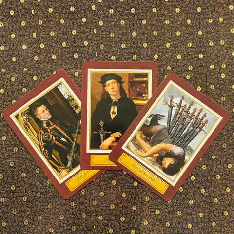 Tarot kortos rinkinys su trijų kortų vėduokle: bordo rėmeliai, viduramžių stiliaus iliustracijos, matoma iš viršaus