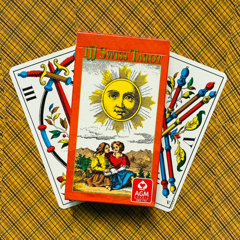 Produkto puslapis — oranžinė JJ Swiss Tarot dėžutė su saulės motyvu, AGM logotipu ir dviem spalvotomis kortomis, nufotografuota iš viršaus