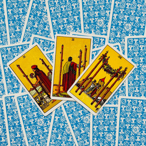 Tarot kortos Rider‑Waite stiliaus, trys atverstos Lazdų (II, III, IV) kortos ant mėlynų raštuotų nugarėlių, skirtos trijų kortų išklotai