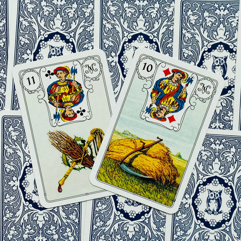 Lenormand kortos, arti dvi atverstos kortos (10 su pjautuvu ir 11 su šluota), matomos kampu, mėlynos nugarėlės su pelėdomis