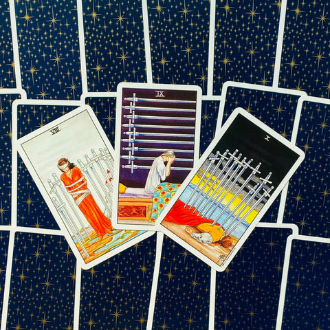 Tarot kortos su tamsiai mėlyna nugarėle ir aukso žvaigždėmis, centrinė trijų atvertų kardų kortų kompozicija (VIII, IX, X).