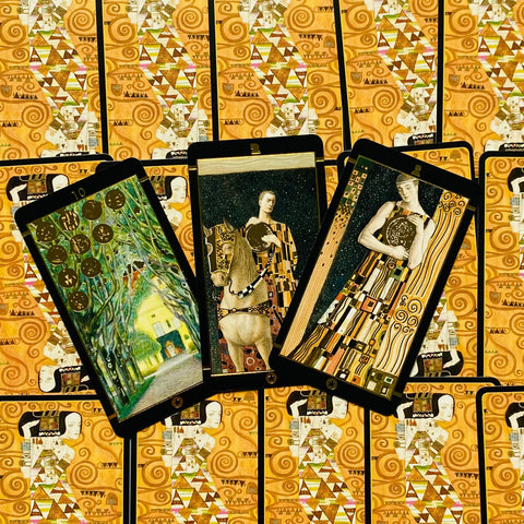 Tarot kortos su Gustav Klimt stiliaus iliustracijomis — trys atverstos kortos virš auksinių nugarėlių, parodyta galerijos variante