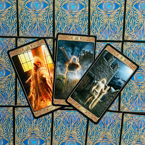 Tarot kortos fanuotos, trys atverstos kortos su auksiniais kraštais ir švytinčiomis figūromis ant mėlyno ornamentuoto nugarėlio