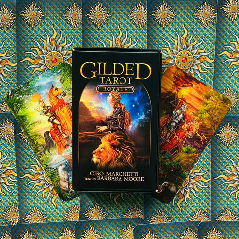 Gilded Tarot kortų rinkinys, juoda dėžutė su liūto motyvu ir dviem išskleistomis kortomis ant ornamentinio fono