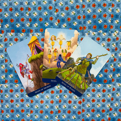 Tarot kortos trijų kortų išklota: Seven of Swords, Seven of Cups ir Seven of Wands ant mėlyno gėlių fono