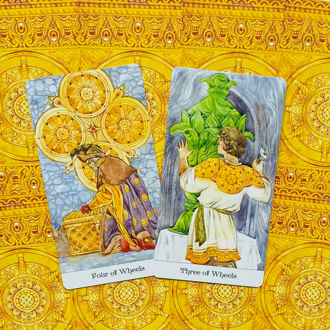 Tarot kortos su aukso ir geltonu ornamentu, atvertos dvi kortos (Three of Wheels ir Four of Wheels) iš komplekto, išdėstytos kampu